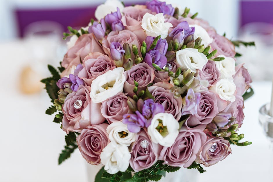 A close-up of a floral bouquet arranged with pale pink roses, white eustoma, and purple freesia flowers, accented with small green buds and lush fern leaves. The bouquet is wrapped in light pink paper