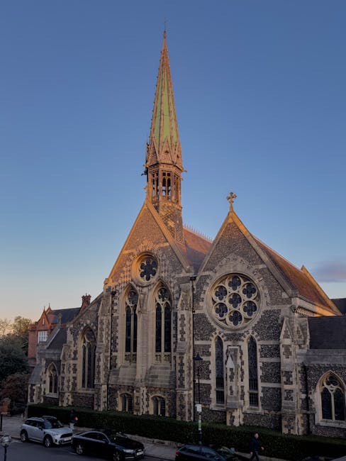 A historic stone church with intricate Gothic-style architecture, featuring pointed arches, large stained glass windows with floral tracery, and a tall, slender spire topped with a cross. The building
