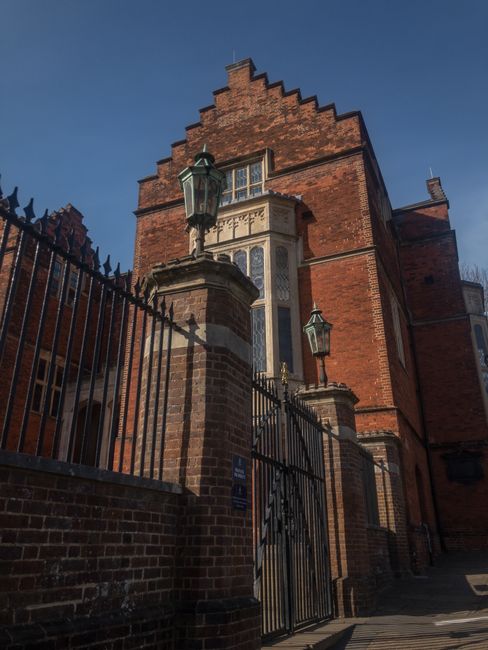 The image shows the exterior of Harrow School, a historic red-brick building with Gothic architectural features, including tall, narrow windows with stone detailing. In the foreground, a wrought iron 