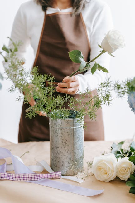 A person wearing a white long-sleeve top and brown apron arranging a simple floral display with green foliage and a single white rose in a tall, cylindrical metal vase. The table is decorated with add