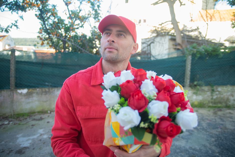 A man dressed in a red cap and matching red jacket stands outdoors holding a vibrant bouquet of fresh roses, predominantly red and white, wrapped in colorful paper. The roses are lush, with open petal