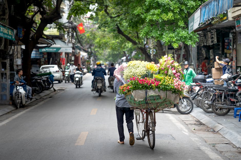 A street scene with a person pushing a bicycle loaded with a large bouquet of fresh flowers, including pink lilies, white hydrangeas, and yellow roses, wrapped in light-colored paper and decorated wit