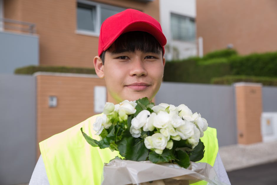 A young delivery person with light skin, wearing a red cap and a fluorescent yellow safety vest, is smiling gently while holding a bouquet of white flowers wrapped in white paper. The bouquet features