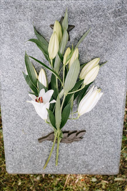 A floral tribute featuring white lilies and white tulips arranged with long, green leaves, secured onto a grey stone memorial. The lilies have large, trumpet-shaped blossoms with a smooth, white textu